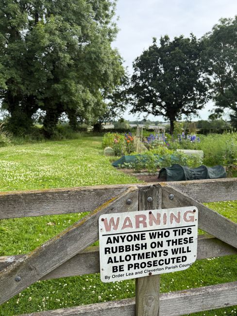 A wooden garden gate with a prominent warning sign attached, reading 'WARNING: Anyone who tips rubbish on these allotments will be prosecuted by order of Lea and Clevendon Parish Council.' Behind the