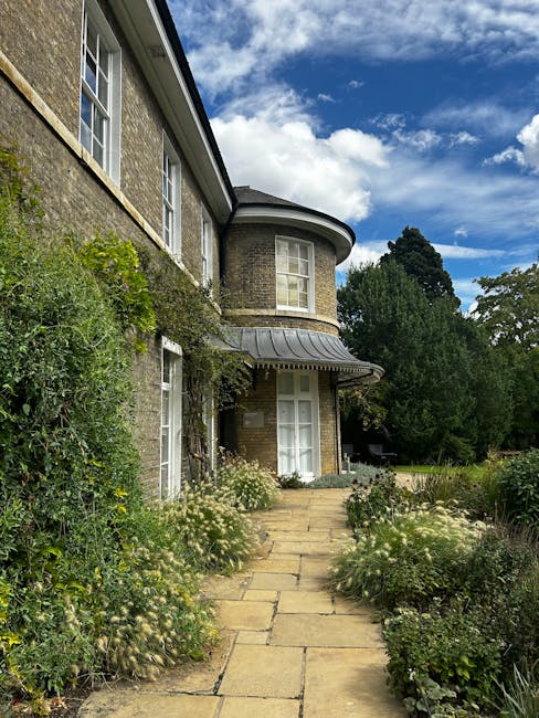 A two-story brick house with a rounded bay window on the upper floor and a smaller bay window on the ground floor, situated at the end of a paved stone pathway. The pathway is flanked by lush, well-ma
